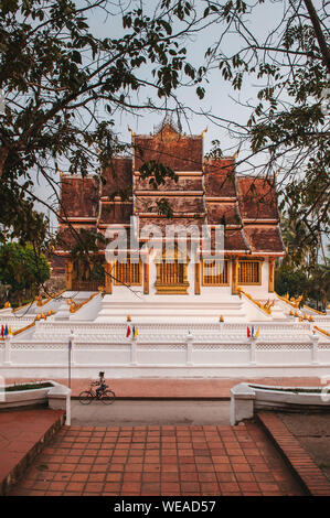 APR 4, 2019 Luang Prabang, Laos - Luang Prabang Royal Palace Museum und Hor Prabang Temple Hall gesehen throuhg Äste mit einem Spaziergang entlang der Straße i Stockfoto