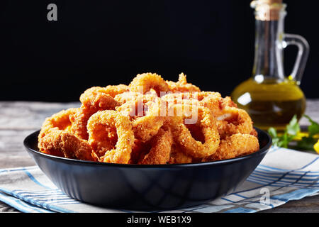 Knusprige calamari Ringe, frittierte panierte Tintenfischringe serviert mit Tomatensauce in einem schwarzen Schüssel auf einen alten Holztisch mit Zutaten auf Schwarz b Stockfoto