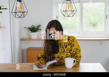 Frau mit Brille mit einem Bleistift auf einem Notebook. Eine weiße Tasse auf einem hölzernen Werkbank in einem Haus. Stockfoto
