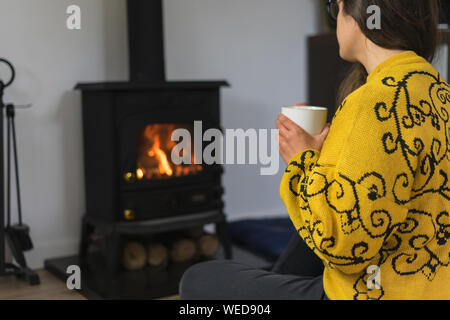 Eine Frau auf dem Boden sitzend genießen Sie Ihr heißes Getränk vor einem Holzofen oder Kamin. Stockfoto