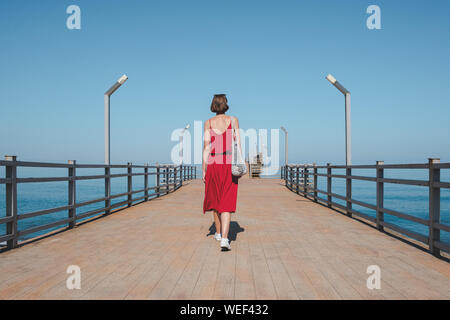 Wandern entlang der Pier mit dem blauen Meer. Die Frau im roten Kleid geht entlang der Kais. Schuß von hinten Stockfoto