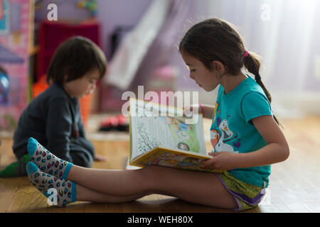 Junge Mädchen lesen Buch, jüngeren Bruder im Hintergrund spielen Stockfoto