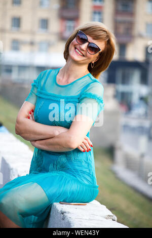 Eine Frau mittleren Alters im Hintergrund sitzt der Stadt auf einem steinernen Brüstung und lächelt in die Kamera, selektiver Fokus Stockfoto