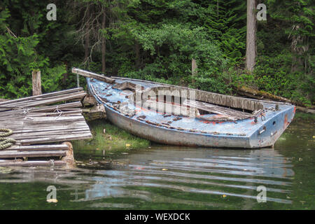 Ein verfallenes, stripped down Alte dayboat sitzt teilweise auf einem grasbewachsenen Ufer neben einem baufälligen Dock in einem bewaldeten Lagune (British Columbia). Stockfoto