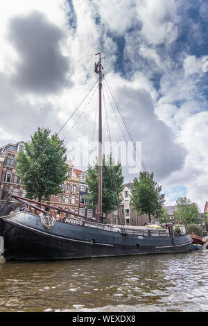 Amsterdam, Niederlande - 1 Juli, 2019: Avance segeln Hausboot am Canal unter schweren cloudscape Leben. Einige grüne Laub. Stockfoto