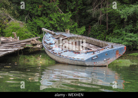 Ein verfallenes, stripped down Alte dayboat liegt teilweise auf dem grasbewachsenen Ufer in einem baufälligen Dock in einer Lagune neben dem Wald (British Columbia). Stockfoto