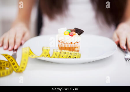 Junge nährende Frau sitzen vor der Platte mit lecker Sahne Torte Kuchen, bei verboten, Lebensmittel mit unglücklich und hungrigen Ausdruck suchen, Studio Stockfoto