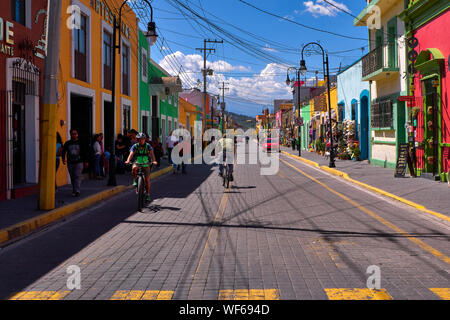 San Pedro Cholula, Mexiko, 30. September 2019 - Schöne Avenida Morelos Straße mit 2 Radfahrer und Touristen an den sonnigen Tag, traditionelle Kleidung und Souvenirläden mit brillanten farbigen Fassaden. Stockfoto