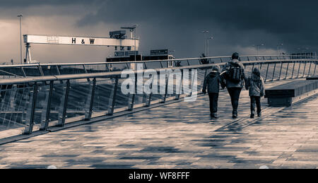 Touristen laufen auf einer Fußgängerbrücke über den Fluss Lagan, während Harland Und Wolffs Kranich Goliath ist in einem Save Our drapiert Yard-Banner Stockfoto