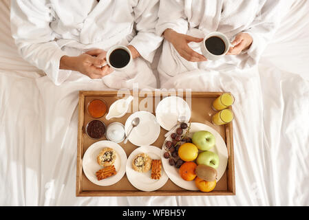 Blick von oben auf die paar tragen weiße Bademäntel im Bett lag. Das Frühstück ist auf das Fach, in das Bett. Zusammen reisen Konzept. Horizontale Schuß Stockfoto