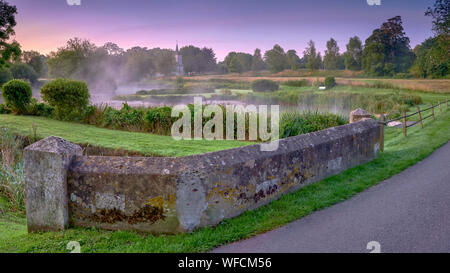 Stoke Charity, Großbritannien - 21.August 2019: Dawn Licht und Nebel auf der alten Mühle Teich im Dorf Stoke Charity mit der Kirche von St. Michael in der Dis Stockfoto