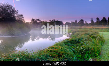 Stoke Charity, Großbritannien - 21.August 2019: Dawn Licht und Nebel auf der alten Mühle Teich im Dorf Stoke Charity mit der Kirche von St. Michael in der Dis Stockfoto