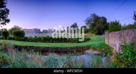 Stoke Charity, Großbritannien - 21.August 2019: Dawn Licht und Nebel auf der alten Mühle Teich im Dorf Stoke Charity mit der Kirche von St. Michael in der Dis Stockfoto