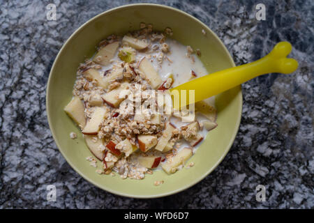 Vorbereitung eines Schweizer Müsli mit frischer Milch, Getreide, trockenen Trauben und Äpfel in einem hellgrünen Schüssel und einem gelben Löffel auf einer Plattform aus Granit in der Küche Stockfoto