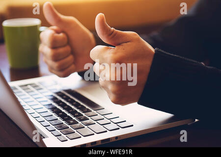 Erfolgreiche gut aussehender Geschäftsmann vor dem Laptop sitzen, Daumen hoch, die im modernen Büro. Erfolgreicher Abschluss der Verhandlungen, gute Arbeitsergebnisse, satis Stockfoto