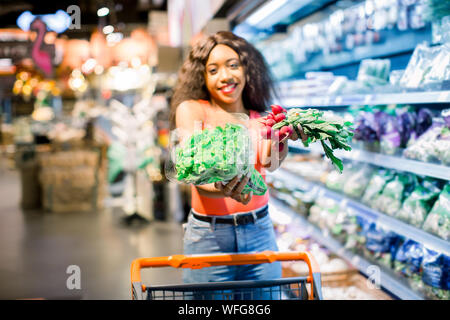 Hübsche Afro junge Frau in Jeans shopping organisches Gemüse und Früchte. Frau mit Katze holding Kopfsalat und Radieschen in ihre Arme. Stockfoto