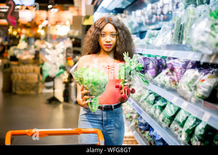 Hübsche Afro junge Frau in Jeans shopping organisches Gemüse und Früchte. Frau mit Katze holding Kopfsalat und Radieschen in ihre Arme. Stockfoto