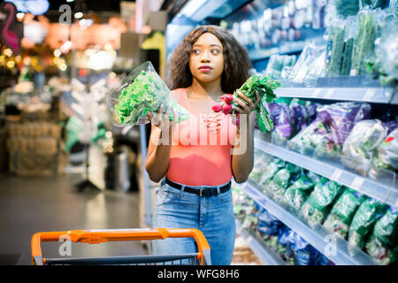 Hübsche Afro junge Frau in Jeans shopping organisches Gemüse und Früchte. Frau mit Katze holding Kopfsalat und Radieschen in ihre Arme. Stockfoto