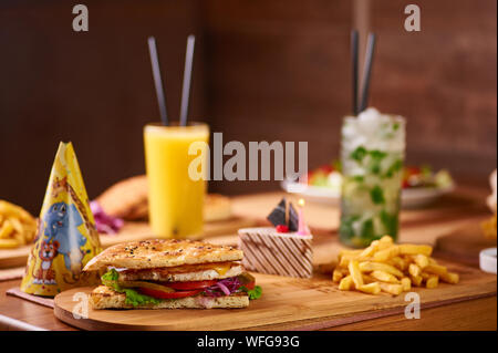 Fast food Tabelle für die Gesellschaft von Freunden serviert. Burger, Pommes und Getränke an Geburtstag serviert. Kinder geburtstag Zusammensetzung von Lebensmitteln, die mit Kuchen mit Stockfoto