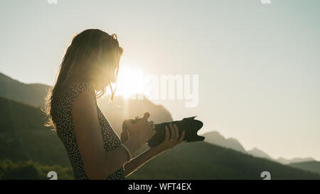 Junge Fotografin an Ihrer DSLR-Kamera einstellen der Eigenschaften, wie sie in der Natur steht, beleuchtet vom Licht der aufgehenden Sonne. Stockfoto