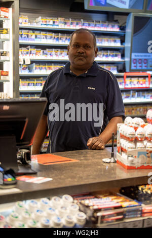 Augsburg, Deutschland. 26 Aug, 2019. Sugendran Veluppillai, Kassierer, ist hinter dem registrieren. Er kam in Deutschland als Asylbewerber, traf seine Frau hier, gründete eine Familie - und jetzt hat er das Geschäft wurde in der Nacht für 18 Jahre in einem großen Augsburg Tankstelle. Der Kassierer Sugendran Veluppillai immer noch begeistert ist über seinen Job an der Kasse der "Esso" Station am Rand der Augsburger Innenstadt. (Dpa: "18 Jahre Tankstelle Nachtschicht - und kein Ende in Sicht") Credit: Lino Mirgeler/dpa/Alamy leben Nachrichten Stockfoto