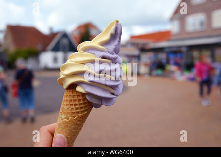 Nahaufnahme einer Hand, die Kegel mit gemischt Gelb und Lila Mango und Berry Softeis mit verschwommenen Stadt Szene im Hintergrund Stockfoto
