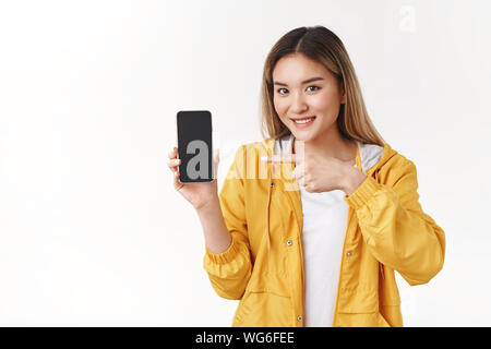 Freundliche nette Angebot asiatische blonde Frau tragen gelbe stilvolle Jacke halten Smartphone anzeigen, der Zeigefinger Telefondisplay breit grinsend Stockfoto