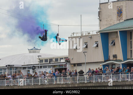 Bournemouth, Dorset, Großbritannien. Mitglieder des Tigers Freefall Parachute Teams stellen fluoreszierende Rauchpfade bereit, während sie auf der Seilbahn fahren, die vom Ende des Piers bis zum Strand beim Bournemouth Air Festival 2019 verläuft. Stockfoto