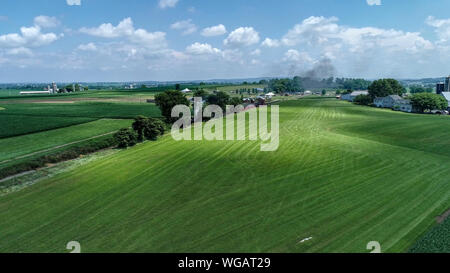 Eine Luftaufnahme von einer Dampflok der Ankunft in Amish Landschaft an einem Sommertag, wie gesehen, die von einer Drohne Stockfoto