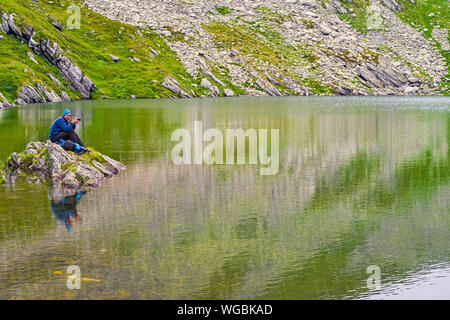 Junger Mann seine Kamera gewinnen an einem klaren Bergsee, stehend auf einem Felsen, Stockfoto