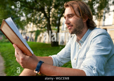 Lächelnd stoppeln Mann hält ein Buch über das Gras in den Campus. Zurück zum Konzept der Schule. Stockfoto