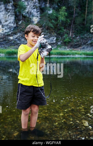 Ein naughty boy, in Shorts und ein gelbes T-Shirt bekleidet, wäscht eine digitale SLR Kamera mit Wasser und Seife von einem Wald Fluss. Wasser in der Stockfoto
