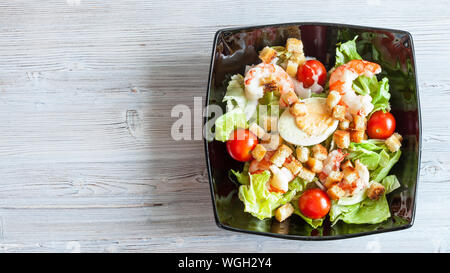 Blick von oben auf die Cesar Salat mit Garnelen in schwarz Schüssel auf grau Holztisch mit Panoramablick copyspace Stockfoto