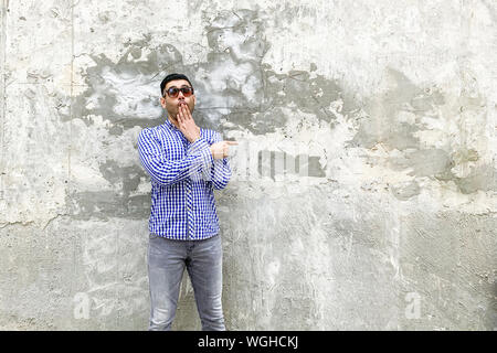 Portrait von schockiert junger Mann in karierten blaues Hemd mit Bart, Sonnenbrille gegen die konkrete graue Wand stehend. Zeigen auf Hintergrund leer copyspace Stockfoto