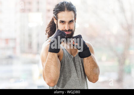 Portrait von zuversichtlich sportlicher junger Erwachsener muskulös Trainer Mann mit lockigem Haar lang stehend mit erhobenen Fäusten schwere Waffen, die im Kampf gegen die Pose, lo Stockfoto