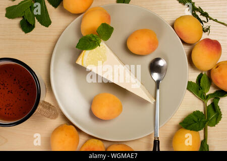 Käsekuchen mit Minze und Zitrone auf einer Platte mit einer Tasse Tee dekoriert ist, wurde von Kräutern und Aprikosen auf hellem Holztisch umgeben Stockfoto