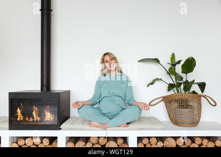 Schwangere Mädchen in grün Sportswear sitzt im Lotussitz Yoga lächelnd in gemütlicher Atmosphäre Stockfoto