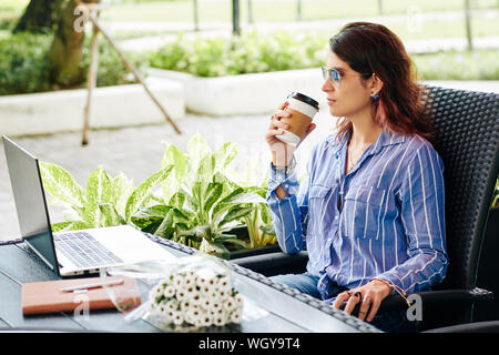 Ernsthafte zuversichtlich Geschäftsfrau in Sonnenbrille im Cafe sitzen vorne geöffnet Laptop und trinken Kaffee Stockfoto