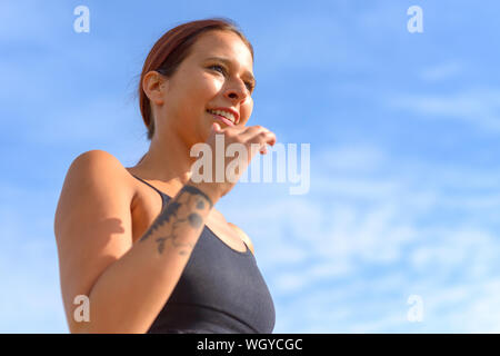 Lächelnde Frau Joggen im Freien im Sommer in der Nähe zu Low Angle View vor blauem Himmel in einem Gesundheit und Fitness Concept Stockfoto