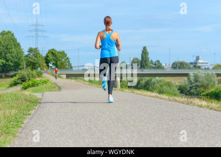 Athletische Frau joggen entlang einer ländlichen Straße neben einem Fluss in einem niedrigen Winkel in voller Länge ansehen von der Rückseite mit Kopie Raum Stockfoto