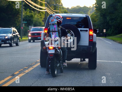 Storrs, CT USA. Aug 2019. Junge Motorradfahrer an einer Kreuzung mit einem Skateboard festgeschnallt auf dem Weg zum Skatepark zu Rucksack. Stockfoto