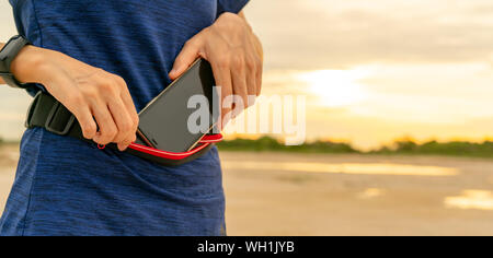 Junge asiatische Frau halten in der Hüfte Smartphone vor der Ausführung von cardio übung am Morgen. Outdoor Training. Läufer und Smart Band tragbare Gerät. Stockfoto