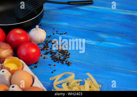 Frühstück. Pfanne, Tomaten, rohe Eier, Zwiebeln, Knoblauch, Gewürze, trockene Teigwaren. Selektive konzentrieren. Kopieren Sie Platz. Auf blau Holz- Hintergrund. Close-up. Stockfoto