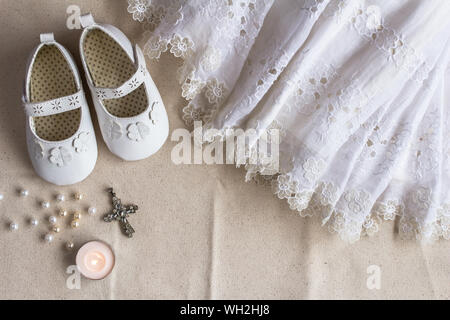 Taufe Hintergrund mit Taufe Kleid, Schuhe, Kerzen und Kristall Kreuz Anhänger auf weißem Leinen Stockfoto