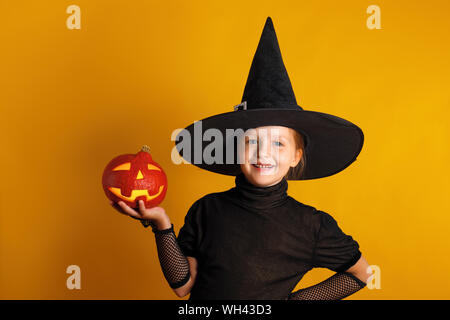 Süße kleine Mädchen in eine Hexe Kostüm hält einen Kürbis jack Lantern auf gelbem Hintergrund. Halloween Feier. Stockfoto