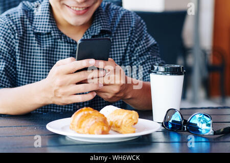 Lächelnden jungen Mann mit Kaffee und Croissants zum Frühstück und Lesen von SMS-Nachrichten auf dem Smartphone Stockfoto