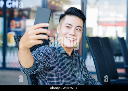 Gutaussehenden jungen vietnamesischen Mann lächelnd bei der Einnahme von selfie auf dem Smartphone im Café im Freien Stockfoto