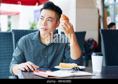 Portrait von Stattlichen nachdenkliche junge vietnamesische Mann essen Gebäck und Kaffee trinken im Café im Freien Stockfoto