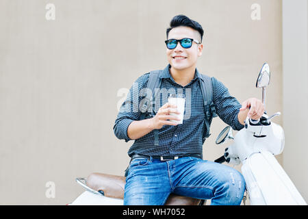 Positive gutaussehenden jungen Mann Sonnenbrille sitzen auf Roller im Freien und trinken lecker Kaffee taje Stockfoto