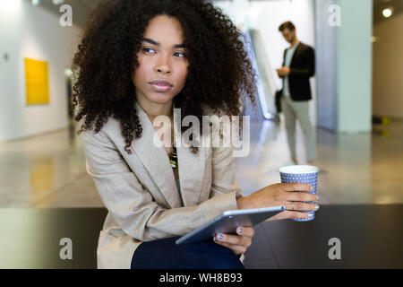 Junge Geschäftsfrau mit Takeaway Kaffee und Tablet-PC in einem Foyer Stockfoto